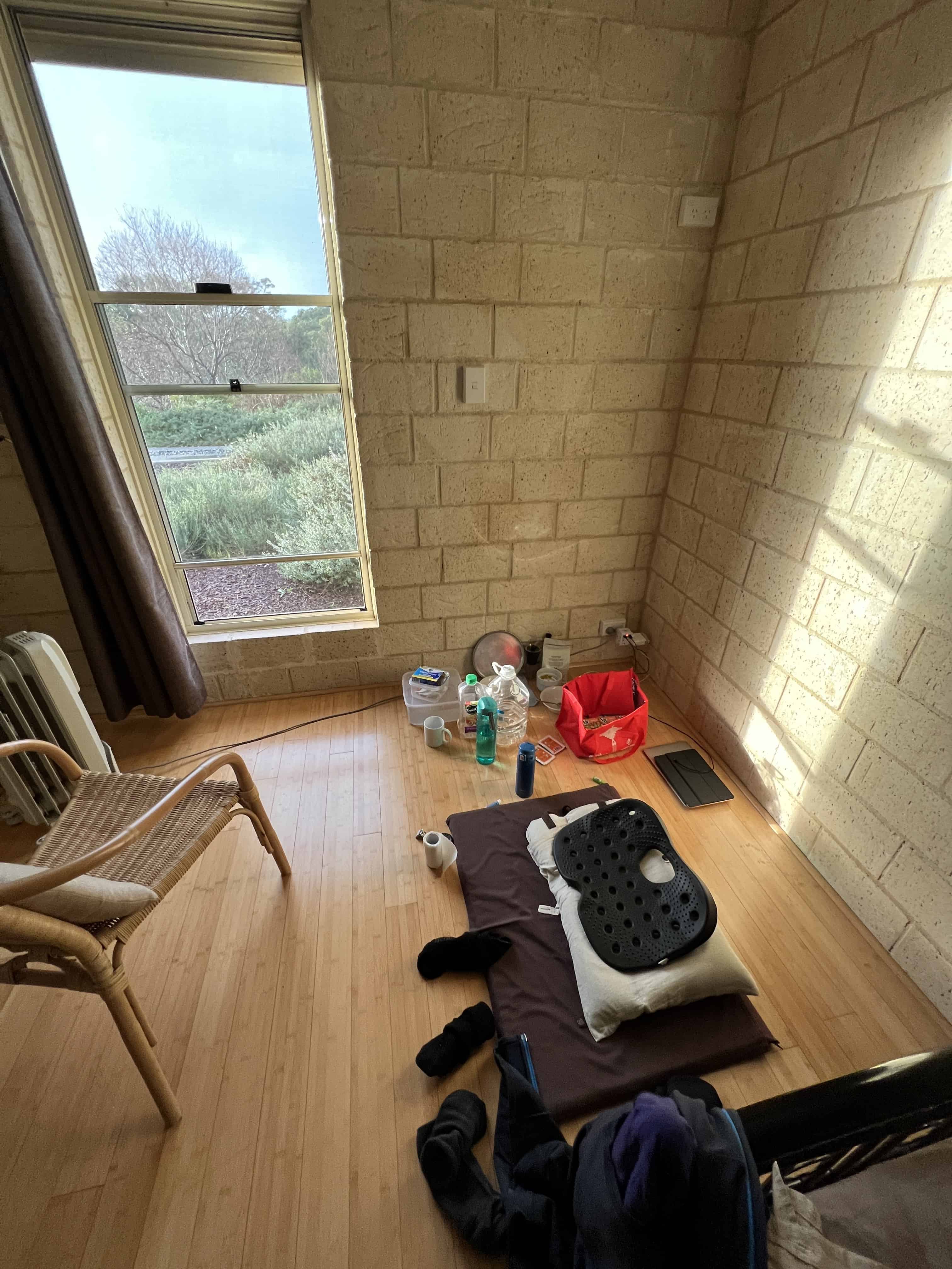 A corner of a room, with a window and sunshine. On the right hand side is a meditation cushion seat. 